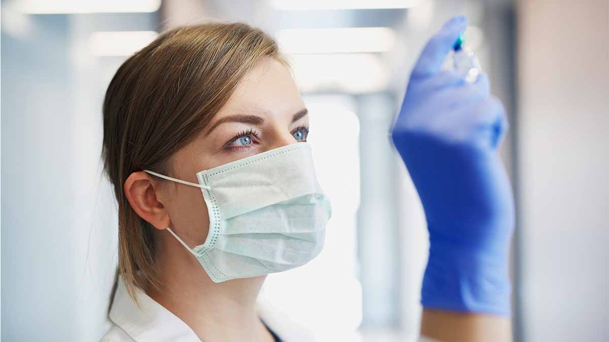 Person wearing gloves and face mask looking at vial they are holding.