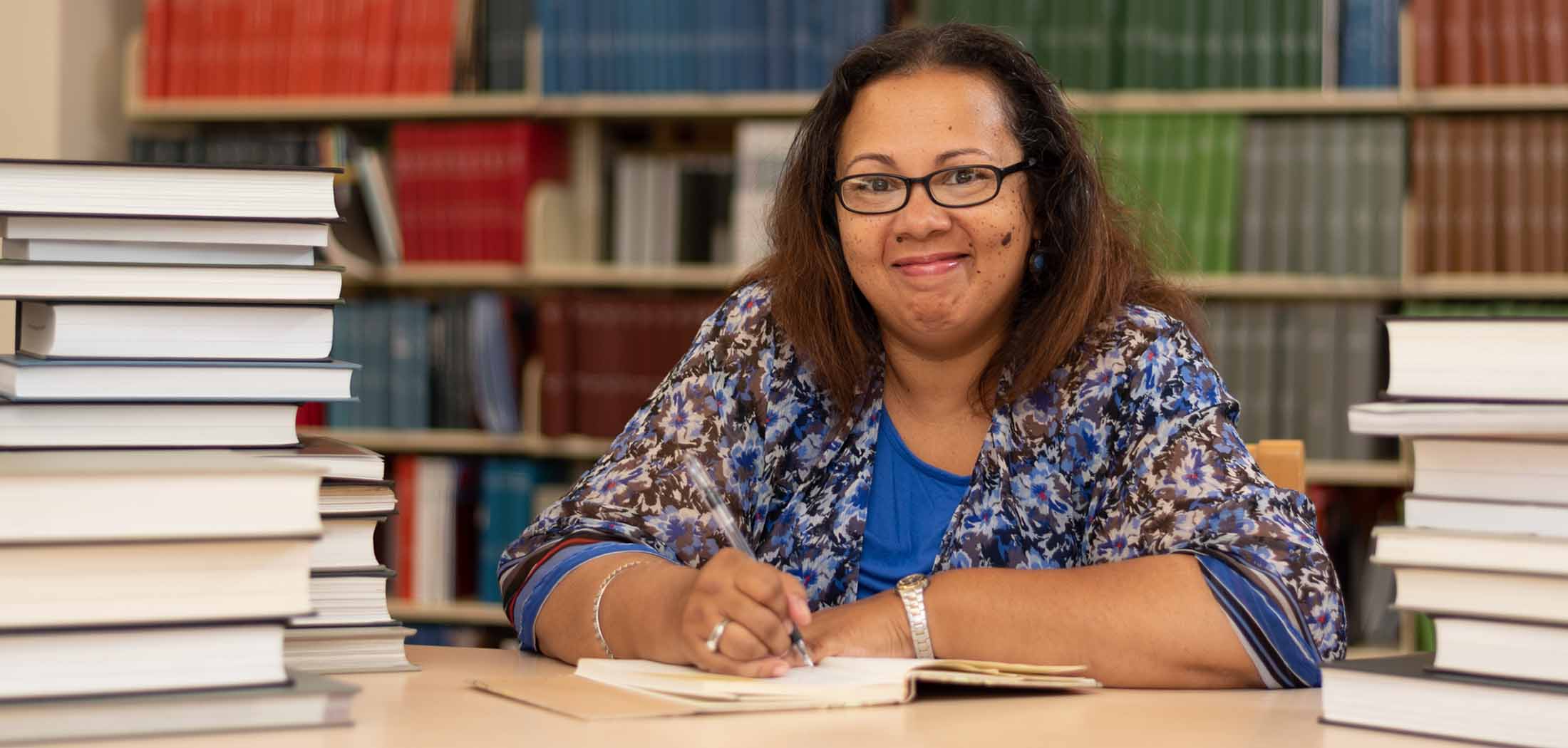 Glynis sitting at a table in a library, holding a pen on a notebook.